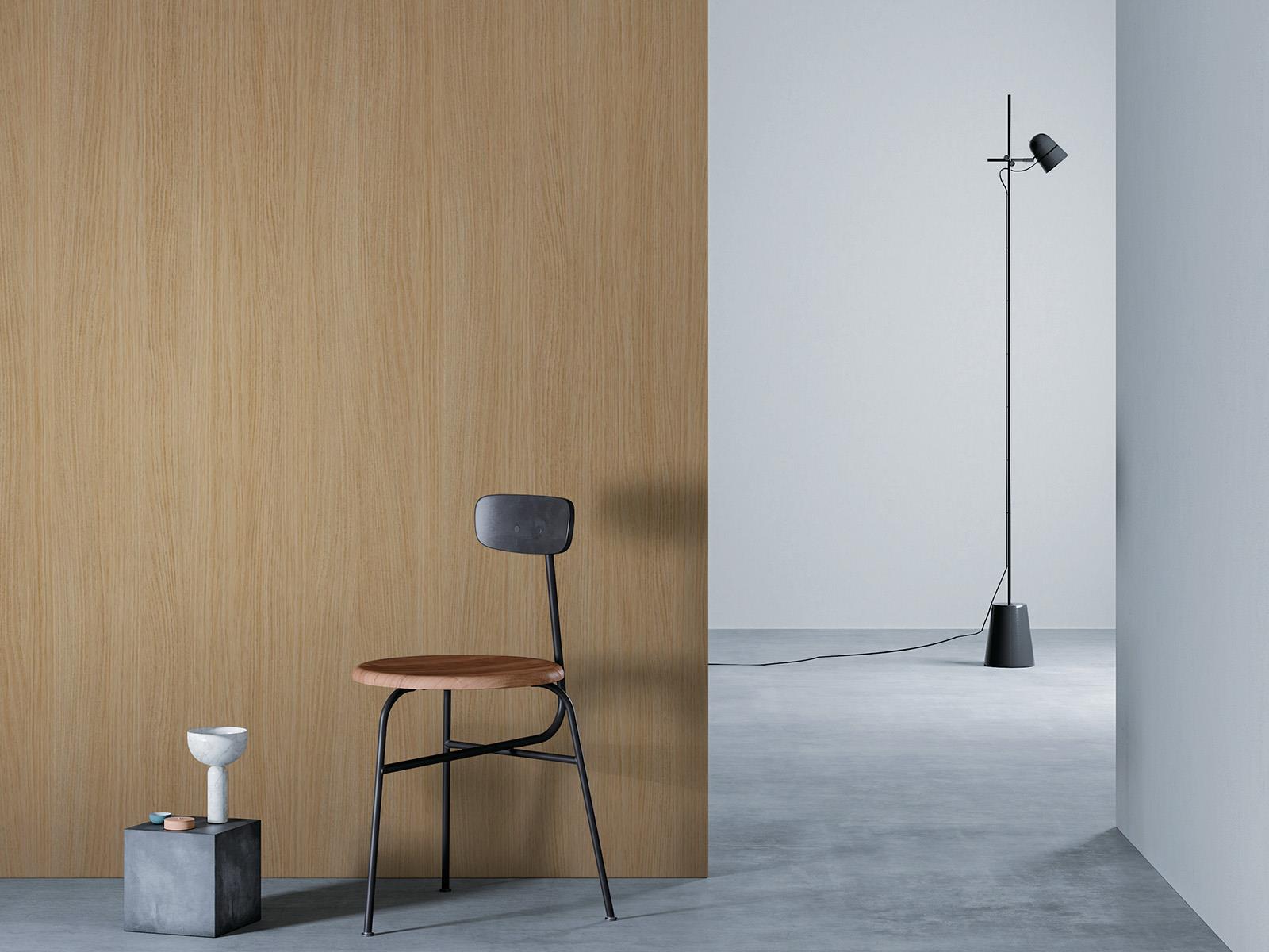 O camera minimalista cu un perete de lemn maro deschis la stanga, o podea gri si un perete alb in dreapta. Un scaun negru cu sezut din lemn este asezat in fata peretelui de lemn, langa un bloc mic de beton cu o vaza alba deasupra. O lampa de podea neagra cu un cablu lung sta in partea dreapta a camerei.