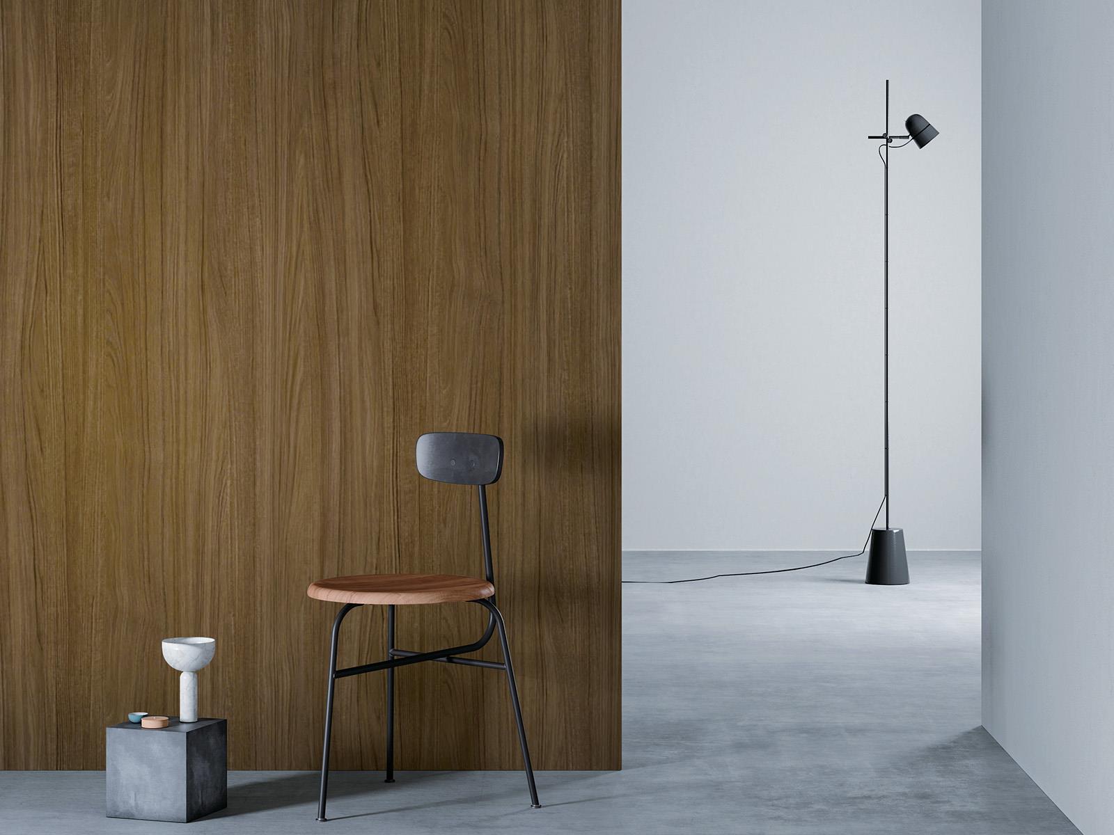 O camera minimalista cu un perete de lemn maro deschis la stanga, o podea gri si un perete alb in dreapta. Un scaun negru cu sezut din lemn este asezat in fata peretelui de lemn, langa un bloc mic de beton cu o vaza alba deasupra. O lampa de podea neagra cu un cablu lung sta in partea dreapta a camerei.