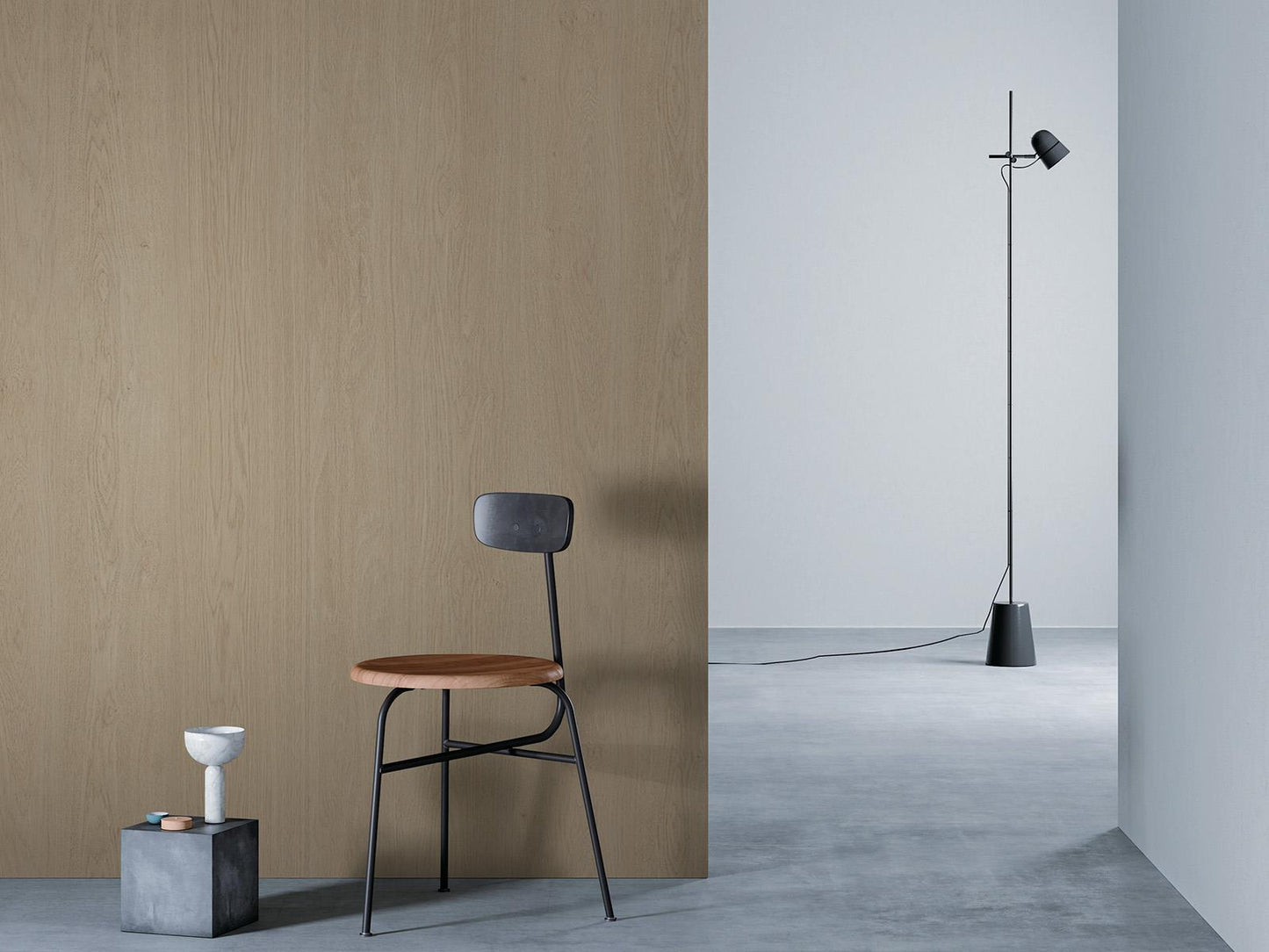 O camera minimalista cu un perete de lemn bej la stanga, o podea gri si un perete alb in dreapta. Un scaun negru cu sezut din lemn este asezat in fata peretelui de lemn, langa un bloc mic de beton cu o vaza alba deasupra. O lampa de podea neagra cu un cablu lung sta in partea dreapta a camerei.