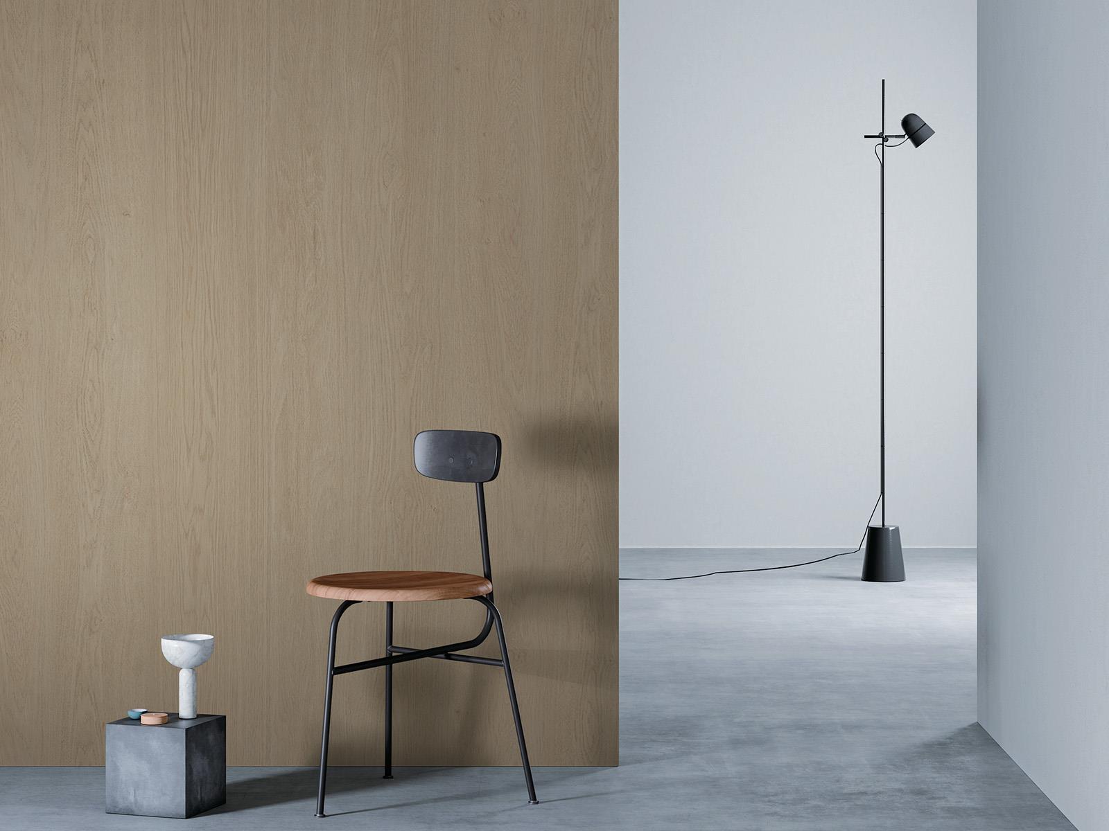 O camera minimalista cu un perete de lemn bej la stanga, o podea gri si un perete alb in dreapta. Un scaun negru cu sezut din lemn este asezat in fata peretelui de lemn, langa un bloc mic de beton cu o vaza alba deasupra. O lampa de podea neagra cu un cablu lung sta in partea dreapta a camerei.