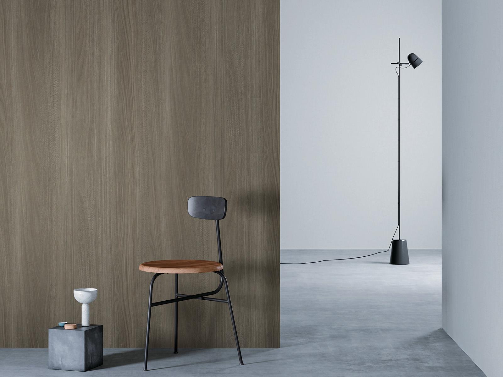 O camera minimalista cu un perete de lemn bej la stanga, o podea gri si un perete alb in dreapta. Un scaun negru cu sezut din lemn este asezat in fata peretelui de lemn, langa un bloc mic de beton cu o vaza alba deasupra. O lampa de podea neagra cu un cablu lung sta in partea dreapta a camerei.