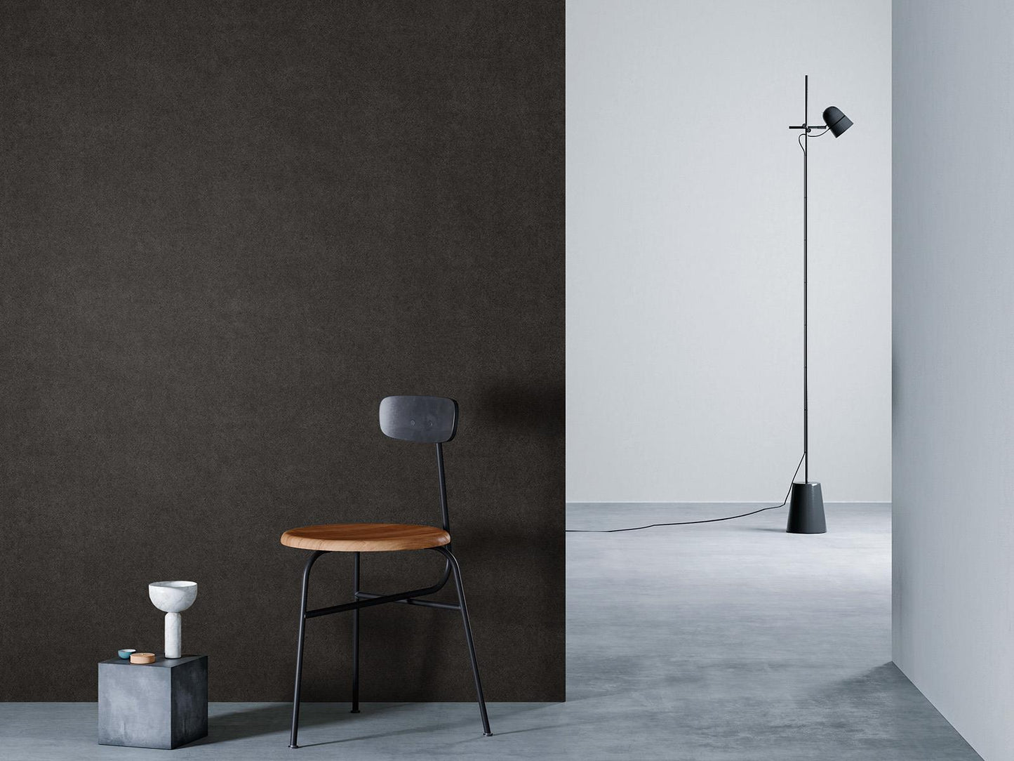O camera minimalista cu un perete maro inchis la stanga, o podea gri si un perete alb in dreapta. Un scaun negru cu sezut din lemn este asezat in fata peretelui de lemn, langa un bloc mic de beton cu o vaza alba deasupra. O lampa de podea neagra cu un cablu lung sta in partea dreapta a camerei.