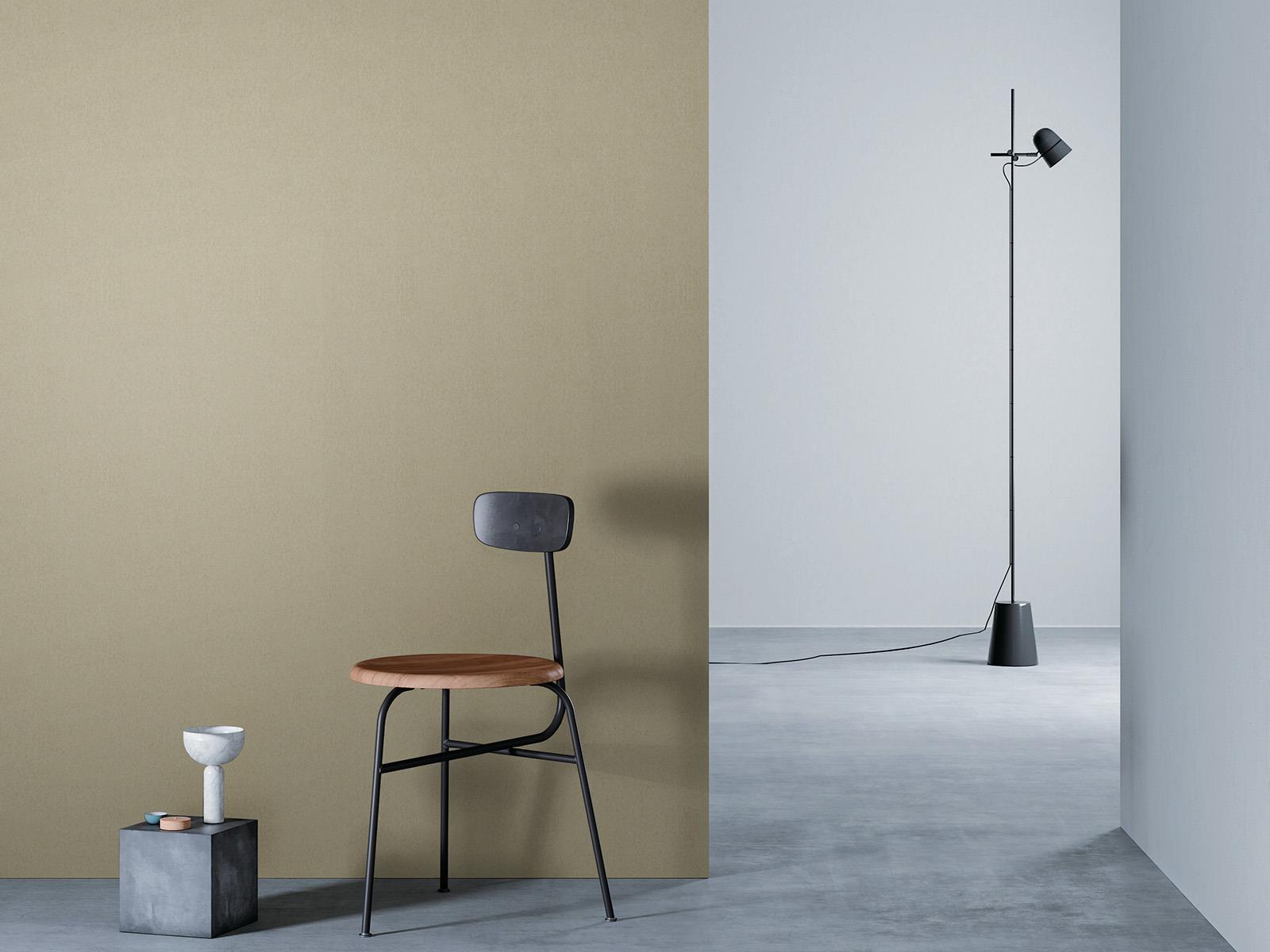 O camera minimalista cu un perete bej la stanga, o podea gri si un perete alb in dreapta. Un scaun negru cu sezut din lemn este asezat in fata peretelui de lemn, langa un bloc mic de beton cu o vaza alba deasupra. O lampa de podea neagra cu un cablu lung sta in partea dreapta a camerei.