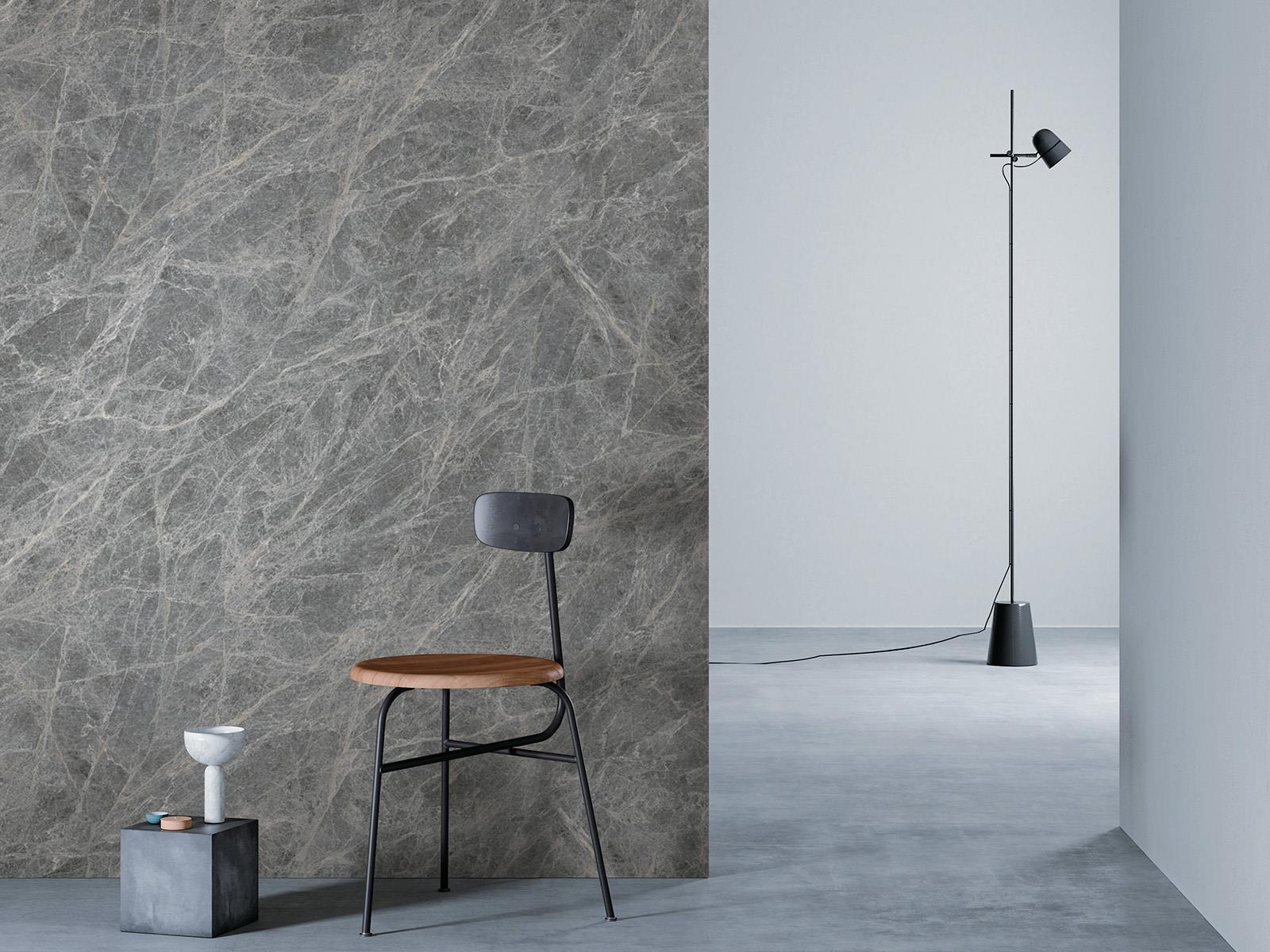 O camera minimalista cu un perete cu faianta gri inchis cu linii gri deschis la stanga, o podea gri si un perete alb in dreapta. Un scaun negru cu sezut din lemn este asezat in fata peretelui de lemn, langa un bloc mic de beton cu o vaza alba deasupra. O lampa de podea neagra cu un cablu lung sta in partea dreapta a camerei.