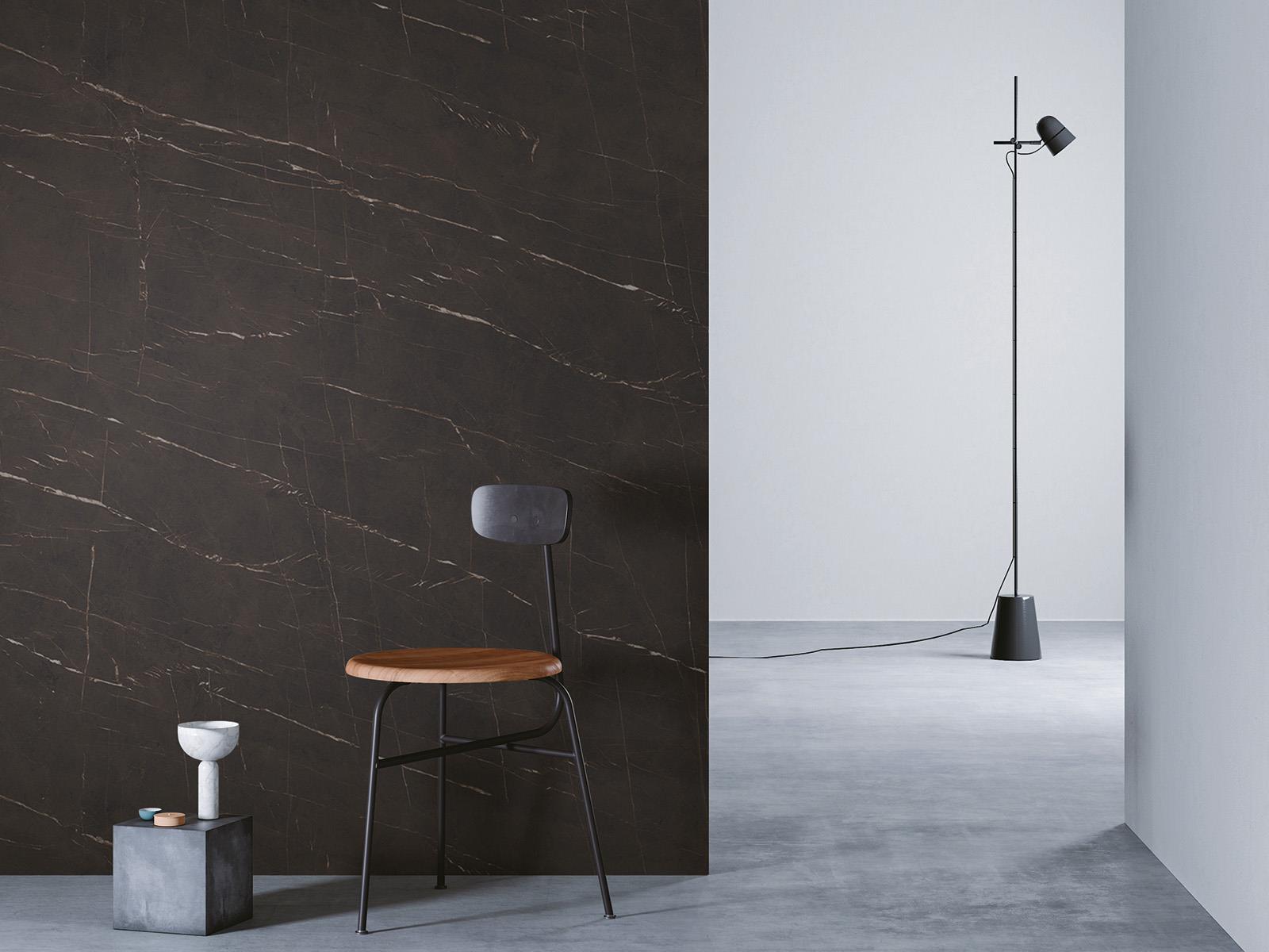 O camera minimalista cu un perete cu faianta maro inchis la stanga, o podea gri si un perete alb in dreapta. Un scaun negru cu sezut din lemn este asezat in fata peretelui de lemn, langa un bloc mic de beton cu o vaza alba deasupra. O lampa de podea neagra cu un cablu lung sta in partea dreapta a camerei.