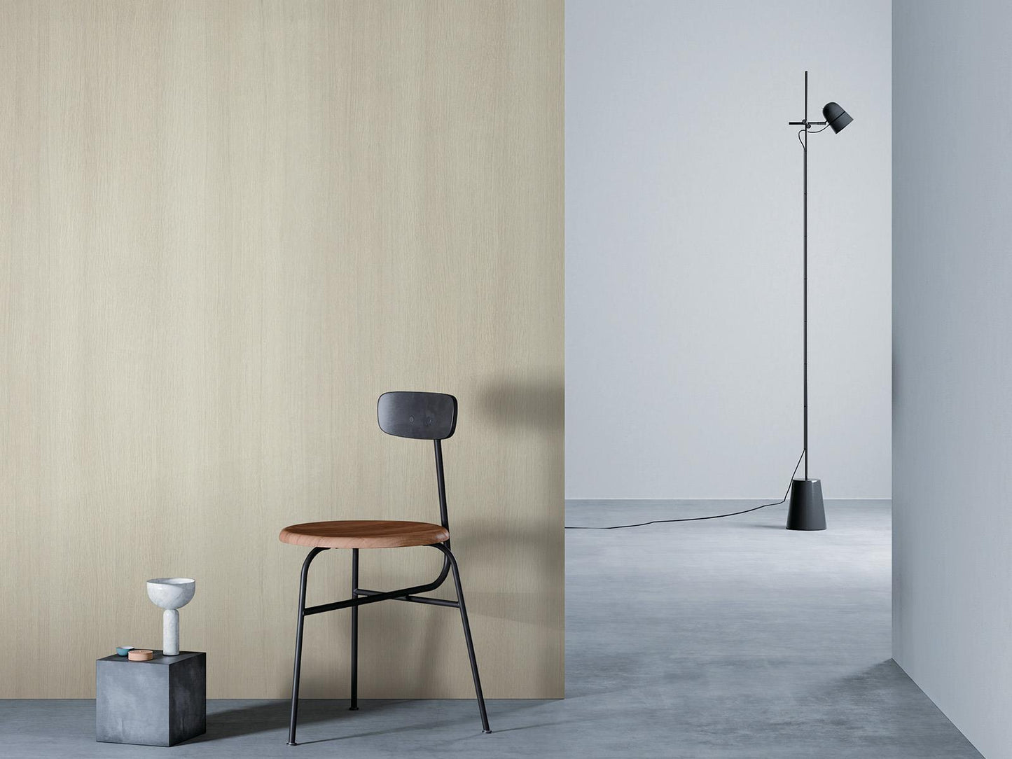 O camera minimalista cu un perete de lemn bej la stanga, o podea gri si un perete alb in dreapta. Un scaun negru cu sezut din lemn este asezat in fata peretelui de lemn, langa un bloc mic de beton cu o vaza alba deasupra. O lampa de podea neagra cu un cablu lung sta in partea dreapta a camerei.