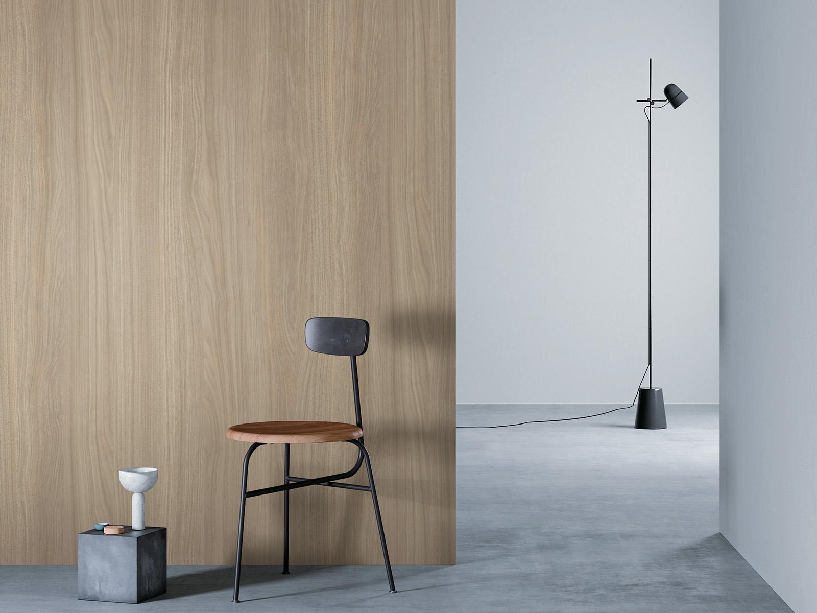 O camera minimalista cu un perete de lemn bej deschis la culoare stanga, o podea gri si un perete alb in dreapta. Un scaun negru cu sezut din lemn este asezat in fata peretelui de lemn, langa un bloc mic de beton cu o vaza alba deasupra. O lampa de podea neagra cu un cablu lung sta in partea dreapta a camerei.
