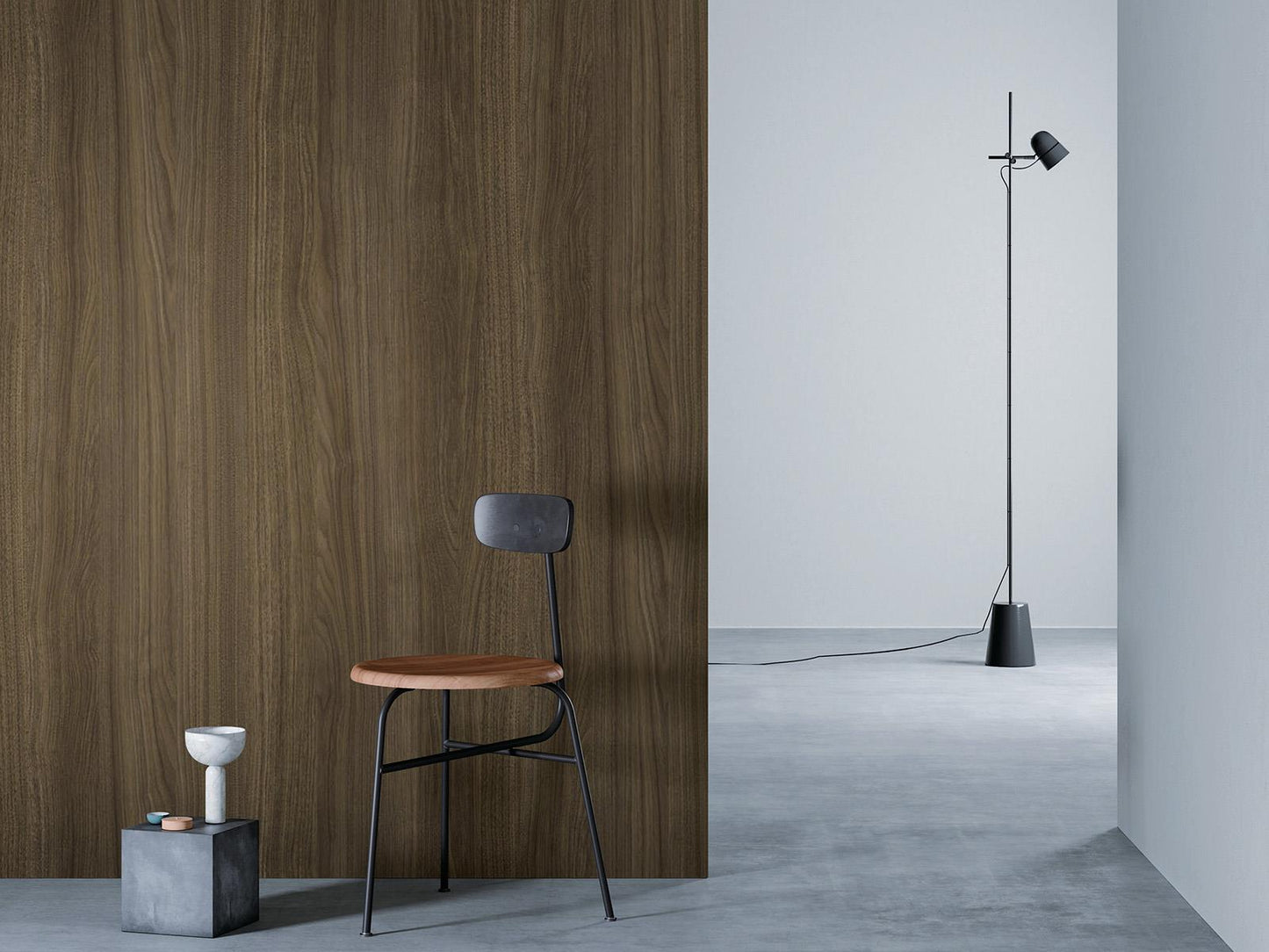O camera minimalista cu un perete de lemn maro deschis la stanga, o podea gri si un perete alb in dreapta. Un scaun negru cu sezut din lemn este asezat in fata peretelui de lemn, langa un bloc mic de beton cu o vaza alba deasupra. O lampa de podea neagra cu un cablu lung sta in partea dreapta a camerei.