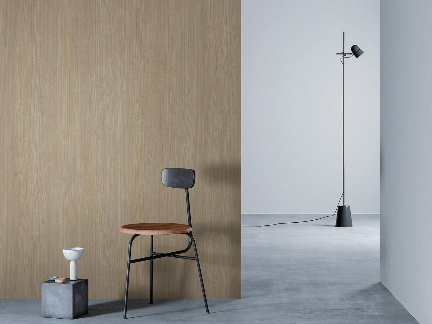 O camera minimalista cu un perete de lemn bej la stanga, o podea gri si un perete alb in dreapta. Un scaun negru cu sezut din lemn este asezat in fata peretelui de lemn, langa un bloc mic de beton cu o vaza alba deasupra. O lampa de podea neagra cu un cablu lung sta in partea dreapta a camerei.