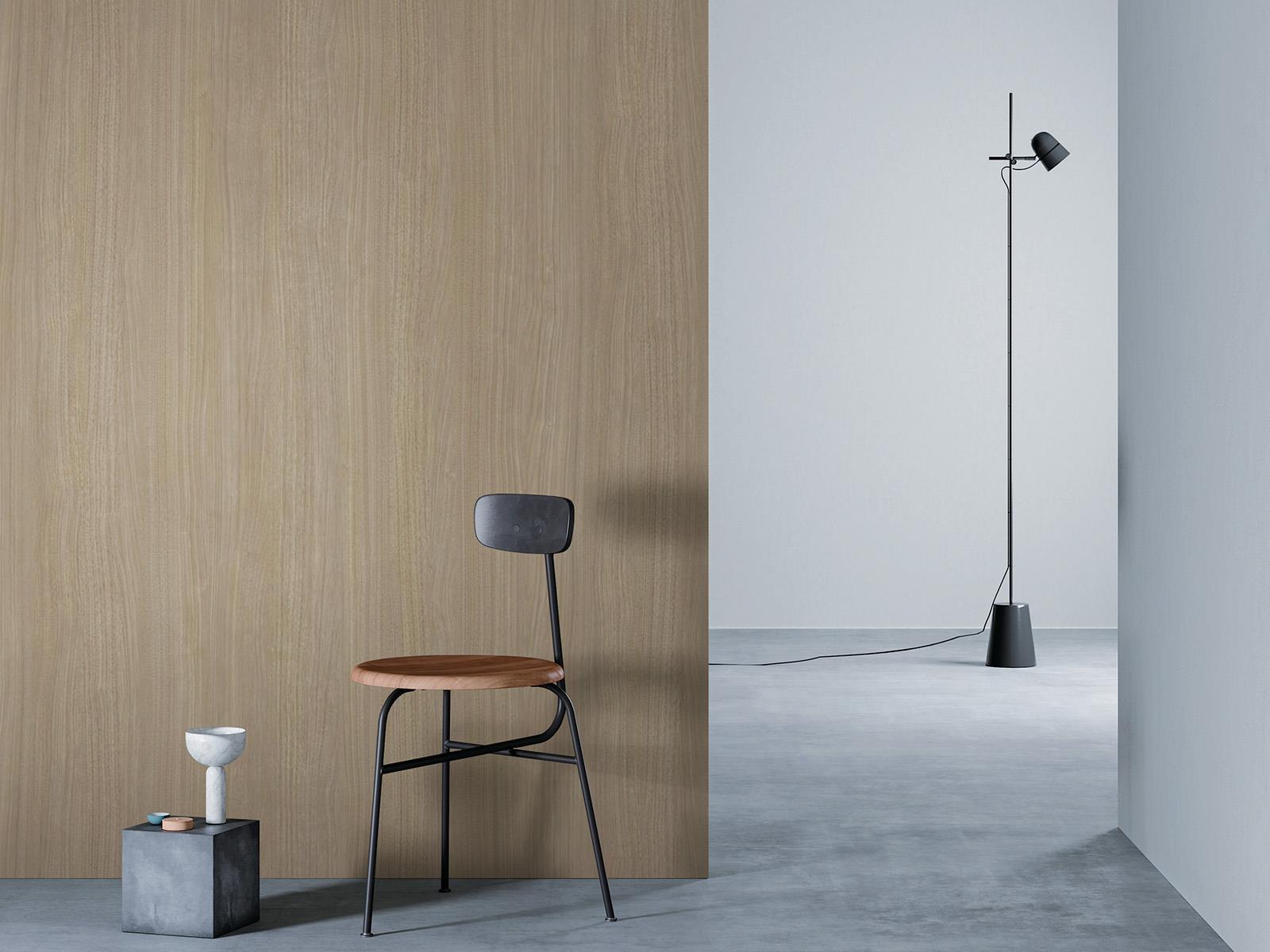 O camera minimalista cu un perete de lemn bej la stanga, o podea gri si un perete alb in dreapta. Un scaun negru cu sezut din lemn este asezat in fata peretelui de lemn, langa un bloc mic de beton cu o vaza alba deasupra. O lampa de podea neagra cu un cablu lung sta in partea dreapta a camerei.