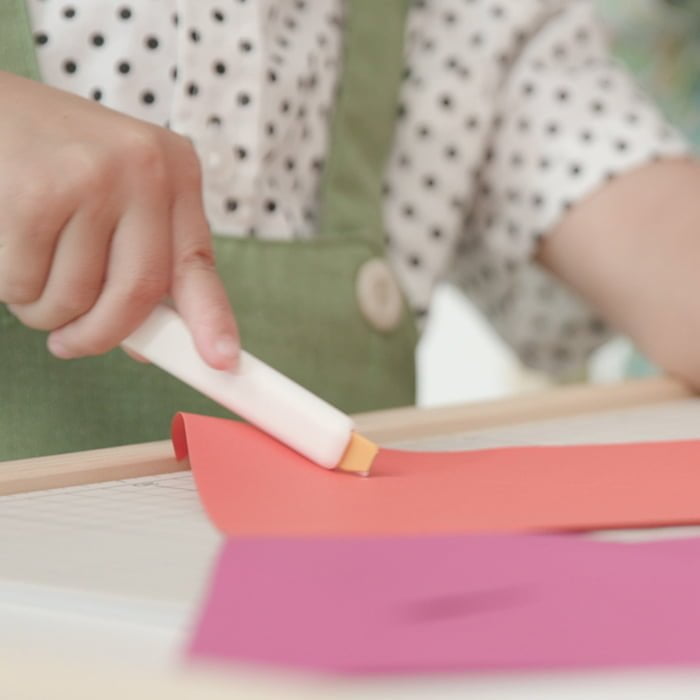 persoana care taie un obiect plan portocaliu cu un cutter alb cu lama protejata