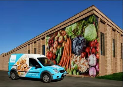 furgoneta albastra de livrare cu reclame pentru alimente sanatoase, parcata in fata unei cladiri cu o pictura murala mare cu fructe si legume.
