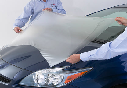 Doua persoane aplica o folie transparenta de protectie a vopselei pe capota unei masini albastre.